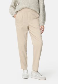 Personne portant un pantalon cargo beige, un pull léger en maille, des chaussettes blanches et des baskets blanches, debout les mains dans les poches.