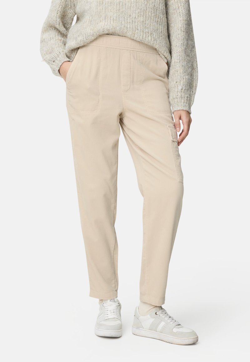 Personne portant un pantalon cargo beige, un pull léger en maille, des chaussettes blanches et des baskets blanches, debout les mains dans les poches.