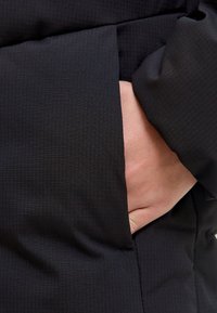 Melna jaka ar teksturētu audumu un smalku režģa rakstu, aprīkota ar sānu kabatu, daļēji noslēpta aiz rokas.