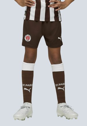 Pantalones cortos marrones de fútbol con el logo del FC St. Pauli, camiseta de rayas blancas, calcetas marrones con el texto "ST. PAULI" y botas de fútbol de color gris claro.
