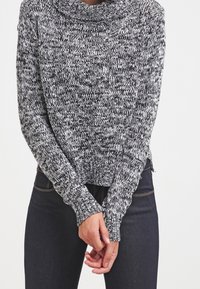 Maglione lavorato a maglia con un motivo mélange bianco e nero, colletto a cowl, maniche lunghe e orlo a costine, con mani visibili vicino alla vita.