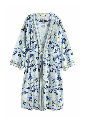 Frau steht barfuß in einem blau-weißen, mit Blumenmuster bedruckten Bademantel, der an der Taille gebunden ist, in einem sanft beleuchteten Wohnzimmer.