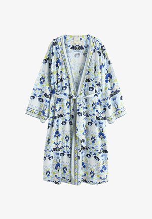 Frau steht barfuß in einem blau-weißen, mit Blumenmuster bedruckten Bademantel, der an der Taille gebunden ist, in einem sanft beleuchteten Wohnzimmer.