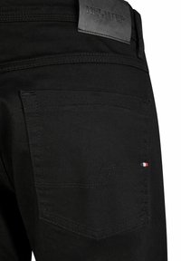 Jean en denim noir avec une poche arrière ornée d'un subtil détail de couture rouge, blanc et bleu, ainsi qu'une étiquette de marque en cuir noir "HECHTER" au-dessus de la taille.