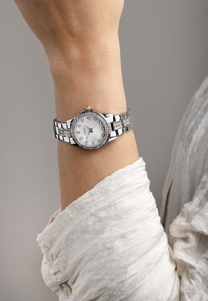 Montre-bracelet en argent avec un cadran blanc rond, une lunette accentuée de cristaux, et un bracelet en maillons métalliques comprenant des maillons polis et texturés.