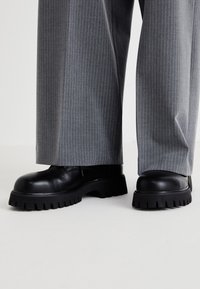 Des chaussures en cuir noir avec une semelle épaisse présentant des rainures profondes, associées à un pantalon large à rayures grises.