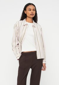 Pruhovaná košeľa v jemnej pastelovej ružovej farbe s zelenými pruhmi, zapínanie na gombíky vpredu, dlhé rukávy, vrstvená cez biely top, kombinovaná s tmavohnedými nohavicami.