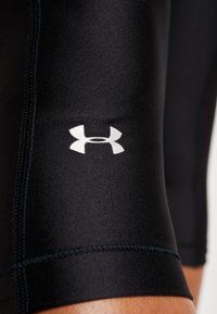 Czarne sportowe szorty o gładkiej fakturze, z białym logo Under Armour na udzie i kontrastującymi szwami.