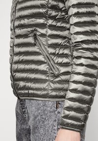 Šedá péřová bunda s prošívanou texturou, boční kapsa na zip a úzké manžety, v kombinaci se světlými džínami.