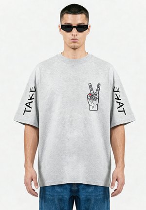 Man met zonnebril en grijs oversized T-shirt met vredeshand-graphic en tekst "Take" op beide mouwen.