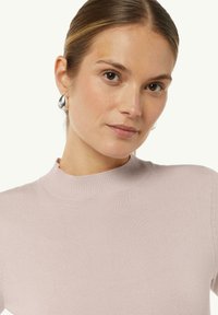 Helles rosa Sweater mit hohem Kragen, geripptem Material und kurzen Ärmeln. Begleitet von einem silbernen Creolenohrring am Modell.