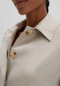 Beige linnen shirt met een kraag, met twee grote, lichtbruine knopen en een gladde, gestructureerde stof. Close-up view benadrukt de details.