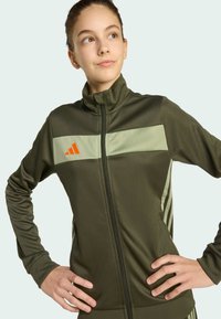 Zelena jakna na zatvarač s svijetlozelenim prugama, s narančastim logotipom na prsima, izrađena od glatkog materijala s dizajnom visokog ovratnika.