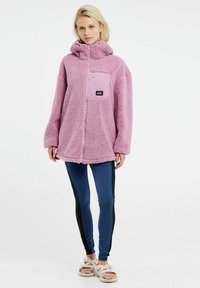 Giacca oversized in pile rosa con cappuccio, tasca frontale e patch con logo, abbinata a leggings blu con strisce laterali nere.