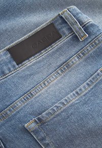 Šviesiai mėlynos spalvos džinsai iš džinso medžiagos su matomu siuvimu, su juodu odiniu antspaudu su užrašu „GABBA“ ir dviem galinėmis kišenėmis.