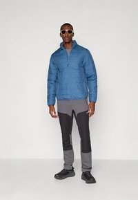 Blå vadderad jacka med blixtlås framtill, bärs över grå och svarta byxor. Modellen står i svarta skor och solglasögon mot en enfärgad bakgrund.