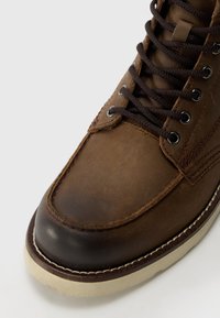 Botín de cuero marrón hasta el tobillo con punta redonda, detalles cosidos, diseño de cordones y una suela de goma crema contrastante. Se incluyen cordones marrones oscuros.