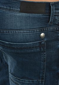 Farkut, joissa on tummansininen väri, näkyvät kontrastipistot ja nahkainen etiketti, jossa lukee "BLEND" vyötärön kohdalla. Metalliset yksityiskohdat korostavat tyyliä.