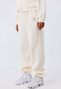 Pantalones de chándal color crema con dobladillo elástico, que presentan un sutil logo plateado. Combinados con zapatillas deportivas en blanco y gris con detalles texturizados.