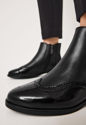 Schwarze Leder-Stiefeletten mit Brogue-Details, glatter Textur, spitzen Zehen und seitlichen Reißverschlüssen. Sie verfügen über einen leichten Absatz und eine robuste Sohle.