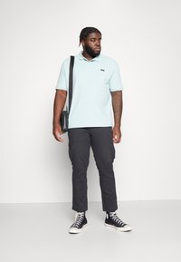 Light blue polo shirt, dark cargo pants, and black high-top sneakers. Model holds a black shoulder bag, with textured hair and neutral backdrop.