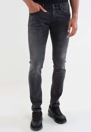 Person wearing faded black slim-fit jeans with hand in pocket and black sneakers, standing on white background.