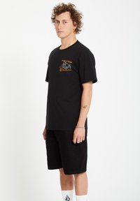 T-shirt en coton noir avec broderie orange "Volcom Workwear" sur la poitrine, assortie à un short noir et des chaussettes blanches. Le modèle se tient de profil.