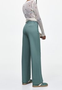 Persona che indossa pantaloni a gamba larga turchesi, sneaker verde e bianco, e un maglione bianco a maglia con fori, in piedi contro uno sfondo bianco uniforme.