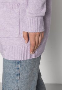 Suéter de punto lila con puños y dobladillo acanalados, que presenta un bolsillo delantero. Combinado con unos jeans de mezclilla azul claro.