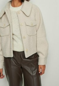 Beżowa, krótkia kurtka z mieszanki wełny z czterema przednimi kieszeniami, dużym kołnierzem i zapięciem na guziki, zestawiona z kremowym, dzianym swetrem i brązowymi spodniami.
