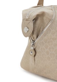 Esquina de una bolsa de tela con patrón beige, con cremallera plateada y tirador redondo de la marca Kipling, que presenta un ribete de cuero cosido y un botón de presión.