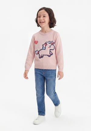 Enfant souriant marchant, portant un pull rose avec un motif de licorne pixellisée et un cœur rouge, un jean bleu et des baskets blanches.