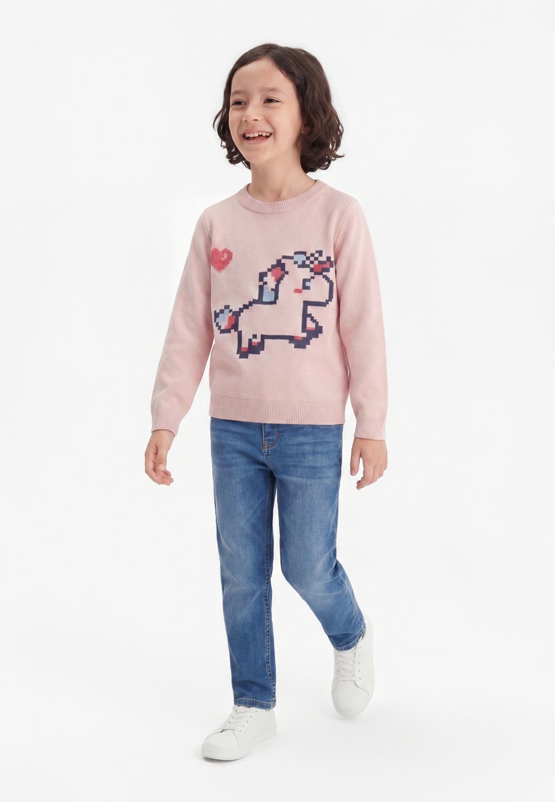 Enfant souriant marchant, portant un pull rose avec un motif de licorne pixellisée et un cœur rouge, un jean bleu et des baskets blanches.