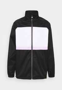 Schwarz-weiße Jacke mit hohem Kragen, Frontreißverschluss und lilafarbenem Horizontalstreifen auf der Brust. Hergestellt aus glattem, leichtem Stoff.