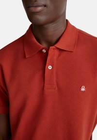 Polo rosso realizzato in cotone textured, con una chiusura a due bottoni, maniche corte e un piccolo logo di un polpo ricamato sul petto.