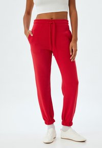 Røde sweatpants lavet af blødt stof, med elastisk talje og snøre, sidelommer og elastiske manchetter ved anklerne.