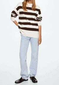 Pull rayé beige et marron, associé à un jean bleu clair et des chaussures noires. Le pull présente une coupe décontractée et des manches retroussées.