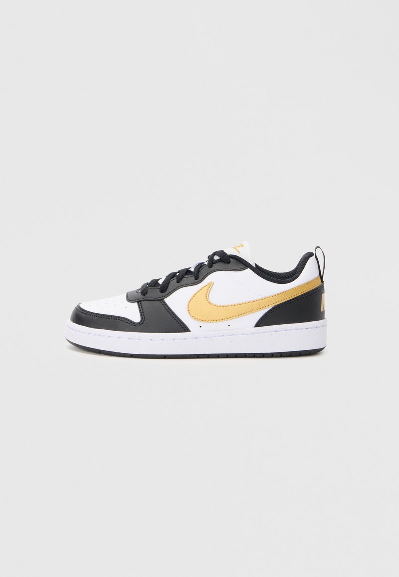 Baskets Nike basse blanches avec des superpositions noires et un swoosh doré, des lacets et un logo au talon, présentées de l'extérieur sur un fond uni.