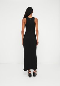 Rochie maxi neagră, fără mâneci, cu un design ajustat, ușor strălucitor și fermoar la spate. Purtată cu sandale negre cu toc gros.