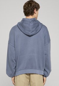 Modrá mikina s kapucňou v uvoľnenom strihu, s prepadnutými ramenami a rebrovanými manžetami. Mäkká textúra a minimálne švy.