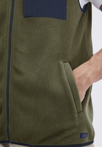 Olivenfarget fleecevest med marineblå detaljer, frontglidelås og to sidelommer. Teksturert stoff med klaffelomme på brystet.