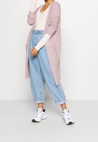 Lång, öppen cardigan i mjuk rosa, lager på lager över en ljusbeige topp. Löst sittande ljusblå jeans och vita sneakers fulländar outfiten.