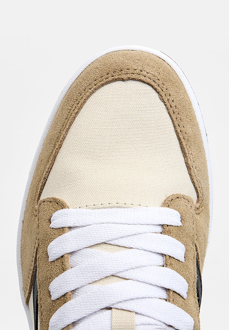 Baskets beige et crème avec une tige en daim et toile, offrant des lacets blancs et une bande latérale noire. Design bas classique avec bout rond.