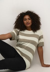 T-Shirt w paski w kolorze kremowym i brązowym, krótki rękaw, luźny krój, miękka tekstura. Połączony z czarnymi spodniami, model siedzi na jasnym tle.