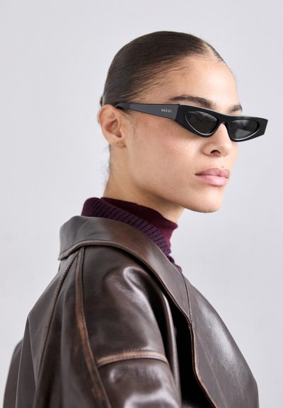Ochelari de soare negri tip cat-eye cu logo pe templu, purtați de un model într-o jachetă de piele maro și un pulover violet, în interior.