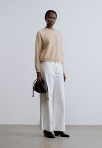 Beżowy, puszysty sweter w połączeniu z białymi spodniami o szerokich nogawkach. Czarna torebka z detalem sznurka, noszona z butami na spiczastym nosku, stojąca na neutralnej powierzchni.