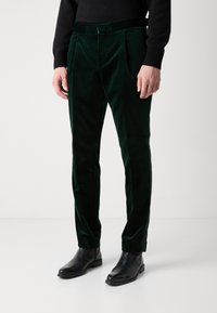 Green velvet trousers featuring two front pleats and a fitted design. Worn with black ankle boots, displaying a soft and textured finish.