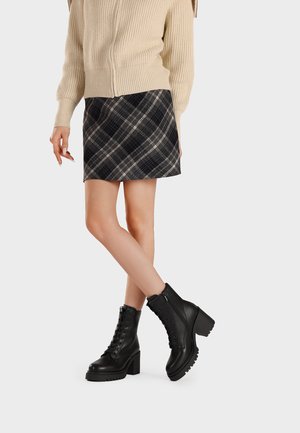Donna che indossa un maglione beige di maglia, gonna a quadri nera e grigia e stivaletti neri stringati con tacchi grossi, in piedi con le gambe incrociate.