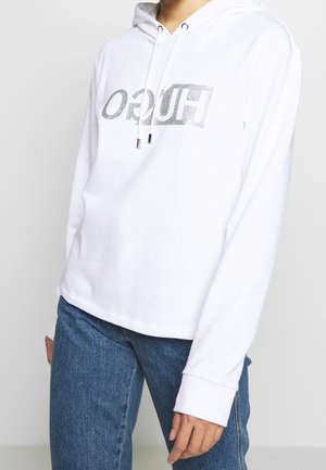 Osoba ubrana w białą bluzę z kapturem z błyszczącym logo "HUGO" i niebieskie dżinsy, stojąca na tle jednolitego jasnego tła.