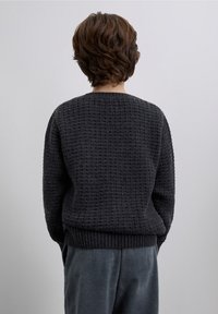 Pull en tricot gris foncé avec un motif texturé, col rond, bordures côtelées aux poignets et à l'ourlet, porté avec un pantalon en velours côtelé. Vue de dos.